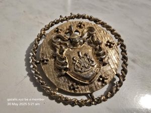 Round metallic ornament with central shield, ornate design, and twisted rope-like border.