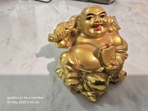Golden laughing Buddha figurine on textured surface with website and date text in corner.