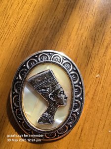 Oval metallic pin with an Egyptian figure profile and decorative border on a wooden surface.