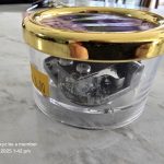 Gold-Lidded Container with Black Object Clear container with gold lid, black metal inside, yellow tag "$3.00" on marble surface.