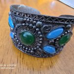 Metallic Cuff Bracelet with Green and Blue Stones Decorative metallic cuff bracelet with green and blue stones on a wooden surface.